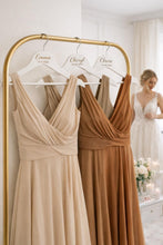 Load image into Gallery viewer, May include: Three bridesmaid dresses in beige and brown tones hang on a gold rail. Each dress has a V-neck and is displayed on a white hanger with a name and date. A bride is visible in the background.
